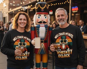 Op de afbeelding: Zwarte sweatshirt met de tekst "WELL CRACK MY NUTS, IT'S WHISKEY O'CLOCK" en een afbeelding van een notenkraker. De afbeelding toont een groot notenkrakerbeeld en twee mensen met whiskeyglazen.