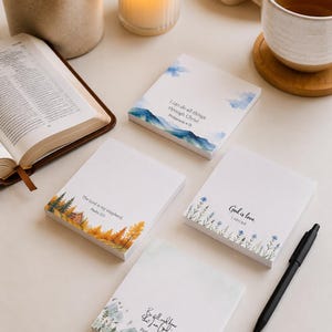 Può includere: Quattro blocchetti di foglietti adesivi quadrati con illustrazioni ad acquerello e versetti biblici. I disegni includono paesaggi montani, alberi autunnali, fiori di campo e scene boschive con i testi I can do all things through Christ, The Lord is my shepherd, God is love e Be still and know that I am God.