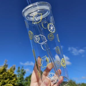 May include: A clear glass tumbler with a lid and straw, adorned with yellow and white lemon designs. The cylindrical glass is held up against a blue sky. The design includes whole and sliced lemons, perfect for summer drinks.