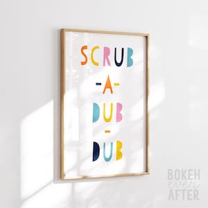 May include: A rectangular wall art print in a light wood frame featuring the text SCRUB-A-DUB-DUB in colorful, rounded, sans-serif letters. The letters are arranged in four lines with orange, yellow, pink, dark blue, and light blue colors.
