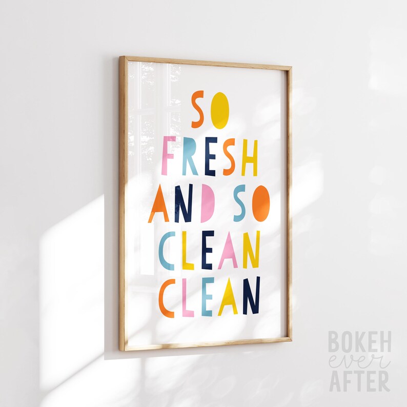 May include: A rectangular wall art print in a light wood frame featuring the text SO FRESH AND SO CLEAN CLEAN in a multi-colored, sans-serif font. The letters are rendered in shades of yellow, orange, pink, light blue, and dark navy blue.