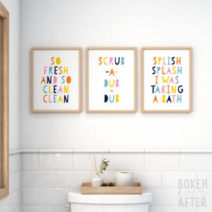 May include: Three rectangular wall art prints in light wood frames. The first print reads SO FRESH AND SO CLEAN CLEAN, the second reads SCRUB-A-DUB-DUB, and the third reads SPLISH SPLASH I WAS TAKING A BATH in colorful, rounded, sans-serif lettering.