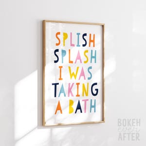 May include: A rectangular wall art print in a light wood frame featuring the colorful text SPLISH SPLASH I WAS TAKING A BATH in shades of orange, yellow, pink, light blue, and dark blue against a white background, displayed in a vertical orientation.