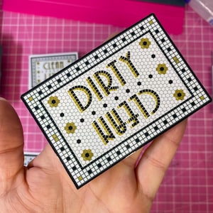 May include: A rectangular sign featuring a mosaic tile pattern in white, black, and gold. The text DIRTY is printed in gold block letters above the word CLEAN, which is also in gold. The design includes a black and white geometric border.