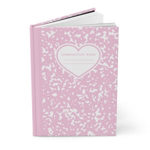 Könnte beinhalten: Ein Notizbuch mit festem Einband und einem hellrosa-weißen Sprenkelmuster. Auf dem Vorderdeckel befindet sich ein weißes, herzförmiges Etikett mit rosa Rand, das den Text COMPOSITION BOOK und zwei horizontale Linien zum Beschriften enthält.