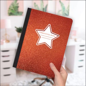 May include: A red glitter composition book with a black spine. A white star-shaped label with the words "COMPOSITION BOOK" is on the front. The notebook is held by a person.