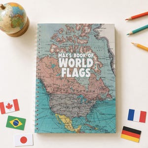 May include: A spiral-bound notebook featuring a vintage-style map of North America on the cover with the text "MAX'S BOOK OF WORLD FLAGS" in white. The notebook is surrounded by a small globe, three colored pencils, and five rectangular flag icons.