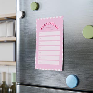 May include: A rectangular magnetic weekly planner with a pink background and white scalloped edges. The text reads WEEKLY PLAN and WEEK/DATE, followed by seven horizontal white rectangular fields labeled MON through SUN for daily scheduling.