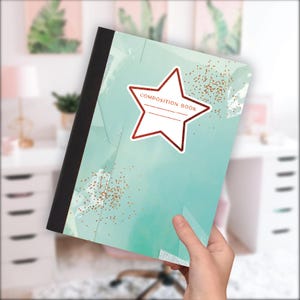 May include: A composition book featuring a teal and white watercolour design, with a star-shaped label that reads "COMPOSITION BOOK". The cover has a black spine and gold glitter accents. The notebook is held at an angle.