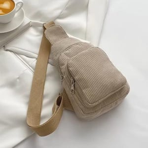 Puede incluir: Un bolso bandolera de pana beige con una sola correa ajustable. El bolso tiene forma rectangular con un bolsillo frontal con cremallera y un compartimento principal. El bolso está hecho de un material de pana suave y acanalado y está diseñado para llevarse cruzado.