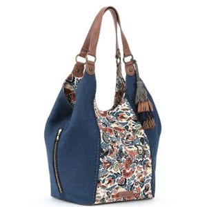 Op de afbeelding: Een blauwe denim hobo-tas met bruine leren riemen en accenten. De tas heeft een stoffen paneel met bloemenpatroon en een zijrits. Een kwast hangt aan de zijkant. De tas is ontworpen voor dagelijks gebruik.