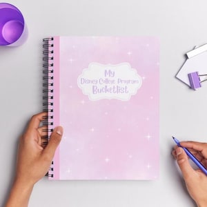 May include: A spiral-bound notebook with a pink cover featuring the text "My Disney College Program Bucketlist" in a white cloud-shaped design. A purple pen and cup are also visible.