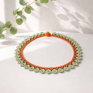May include: A beaded necklace with a floral pattern. The necklace has an orange base with green and red details. The beaded design forms a scalloped edge. The necklace is displayed on a white surface.