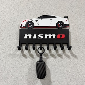 May include: A black key holder with six hooks, featuring a white sports car design with red accents and the word "NISMO" in white and red. A key with a carbon fiber case hangs from the bottom.