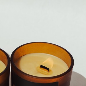 K&ouml;nnte beinhalten: Nahaufnahme einer brennenden Kerze in einem braunen Glasbeh&auml;lter. Die Kerze hat einen Holzdocht und cremefarbenes Wachs. Der Beh&auml;lter hat ein schwarzes Etikett mit wei&szlig;em Text "Olive & Ember Co".