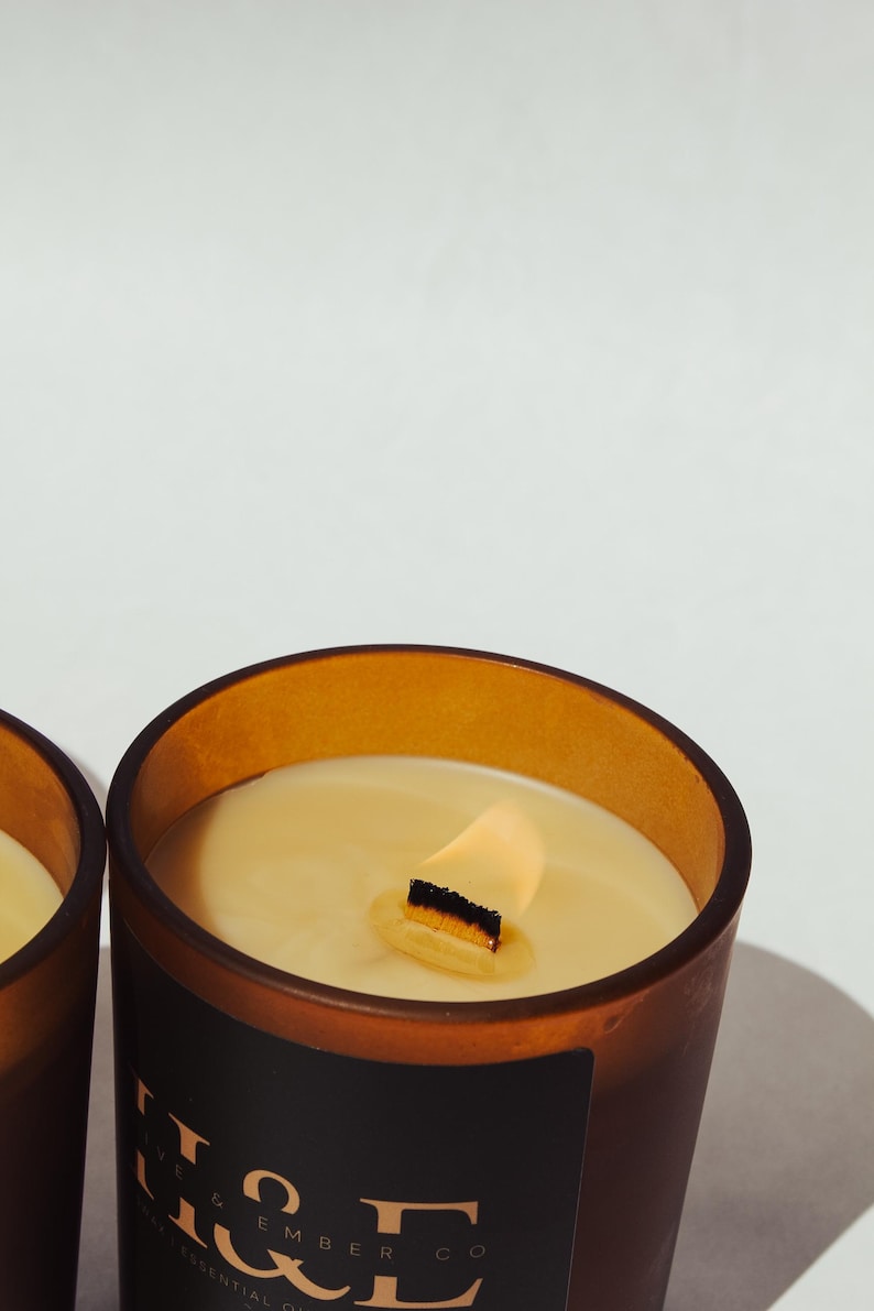 K&ouml;nnte beinhalten: Nahaufnahme einer brennenden Kerze in einem braunen Glasbeh&auml;lter. Die Kerze hat einen Holzdocht und cremefarbenes Wachs. Der Beh&auml;lter hat ein schwarzes Etikett mit wei&szlig;em Text "Olive & Ember Co".