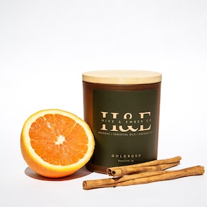 K&ouml;nnte beinhalten: Eine braune Kerze mit Holzdeckel, einer aufgeschnittenen Orange und Zimtstangen. Die Kerze hat ein dunkelgr&uuml;nes Etikett mit dem Text "HIVE & EMBER CO" und "GOLDRUSH". Die Kerze ist 226,7 g.