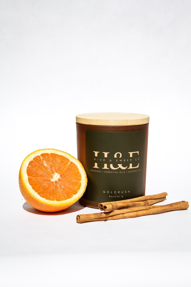 K&ouml;nnte beinhalten: Eine braune Kerze mit Holzdeckel, einer aufgeschnittenen Orange und Zimtstangen. Die Kerze hat ein dunkelgr&uuml;nes Etikett mit dem Text "HIVE & EMBER CO" und "GOLDRUSH". Die Kerze ist 226,7 g.