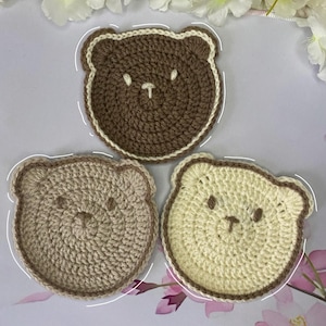 May include: Three handmade crochet bear coasters in shades of brown and cream. Each coaster features a bear face design with a contrasting border. The coasters are round and appear to be made of soft yarn.