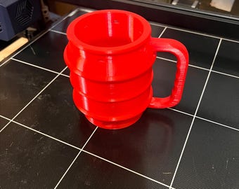 Può includere: Una tazza rossa brillante, stampata in 3D, con un manico. La tazza ha una forma cilindrica con scanalature orizzontali. È appoggiata su una superficie nera con un motivo a griglia, probabilmente un piano di stampa 3D. La tazza è l'elemento principale dell'immagine.