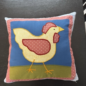 May include: A square throw pillow featuring a central applique of a yellow chicken with a red patterned wing, comb, and wattle. The chicken stands on a green patch against a blue background, framed by a pink border with white hearts and a light blue edge.