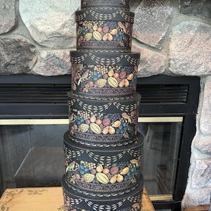 May include: A stack of six round, black nesting boxes with lids. Each box features a painted design of a basket filled with colourful fruits, including pears, grapes, and berries, accented by small birds and a repeating leaf border pattern.