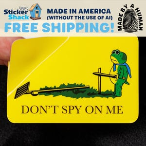 May include: A rectangular yellow sticker featuring a green frog wearing a blue cape and holding a sword. The frog stands next to a dark green patch of grass and a black rake. The text DON'T SPY ON ME is printed in black at the bottom of the sticker.
