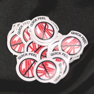 Puede incluir: Un mont&oacute;n de pegatinas blancas y circulares con un dise&ntilde;o gr&aacute;fico rojo y negro. El dise&ntilde;o presenta las letras "WR" tachadas con una l&iacute;nea roja, rodeadas por un c&iacute;rculo rojo. Las palabras "QUICK PEEL" est&aacute;n impresas en negro alrededor del borde de cada pegatina.