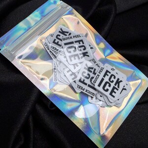 K&ouml;nnte beinhalten: Eine transparente, irisierende Plastikt&uuml;te mit mehreren wei&szlig;en Aufklebern. Jeder Aufkleber tr&auml;gt den Text "FCK ICE QUICK PEEL" in schwarzer, fetter Schrift. Die T&uuml;te ist versiegelt und liegt auf einer schwarzen Oberfl&auml;che.