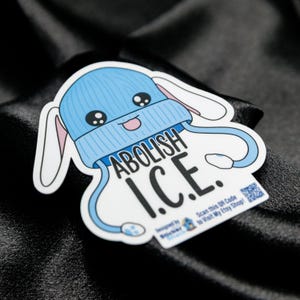 May include: A sticker featuring a cartoon bunny wearing a light blue beanie with the words "ABOLISH I.C.E." The sticker has a white border and a QR code. The bunny has large eyes and a smiling mouth.