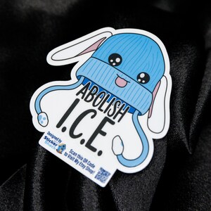 May include: A white sticker with a cartoon design of a blue beanie with bunny ears and the text "ABOLISH I.C.E.". The sticker also includes the text "Designed by Sticker Shack" and a QR code.