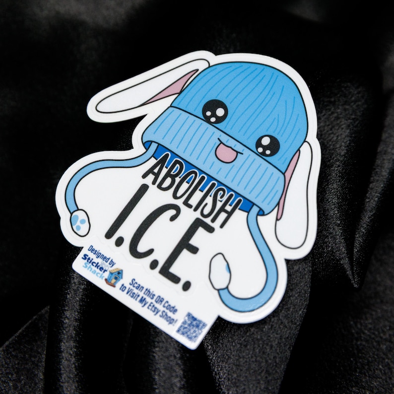 May include: A white sticker with a cartoon design of a blue beanie with bunny ears and the text "ABOLISH I.C.E.". The sticker also includes the text "Designed by Sticker Shack" and a QR code.
