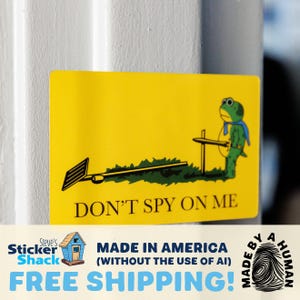 May include: A rectangular yellow sticker featuring a green frog wearing a blue cape and holding a sword. The sticker displays a graphic of a rake lying on the ground and includes the black text DON'T SPY ON ME printed at the bottom of the design.