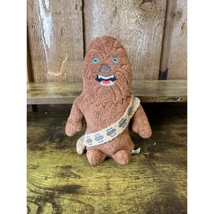 May include: A brown plush toy resembling a Wookiee character, featuring blue eyes, a stitched mouth, and a bandolier with blue and beige accents. The toy is made of soft, textured material.