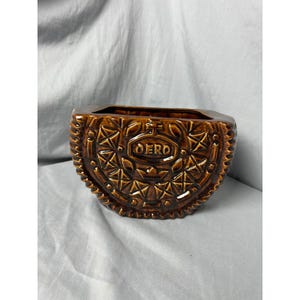 May include: A brown ceramic planter shaped like an Oreo cookie. The planter has a square opening and is decorated with the word "OERO" in the center. The cookie design includes star and geometric patterns.