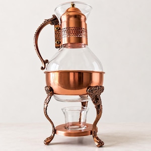 May include: A glass carafe with a copper-colored metal base, lid, and handle. The carafe features an ornate, filigree-patterned copper band around the neck and rests on a stand with three decorative legs, including a small glass cup underneath.