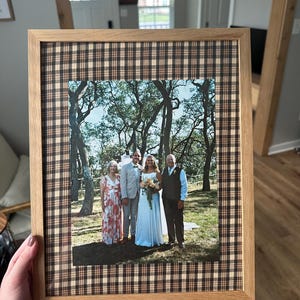 May include: A framed photograph of a wedding party. The frame is light brown wood with a plaid patterned border. The photograph shows a bride in a white dress, a groom, and two other adults. The background is a natural outdoor setting.