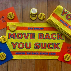 May include: A yellow rectangular towel with orange text reading THE SWAMP MOVE BACK YOU SUCK MOVIN 'EM BACK SINCE 1930, accompanied by several yellow circular buttons with the same text, and orange and blue cards featuring a small graphic.