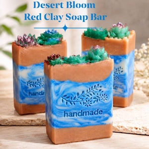 Könnte beinhalten: Drei rechteckige Seifenstücke mit der Aufschrift Desert Bloom Red Clay Soap Bar. Jedes Stück hat eine hellbraune Basis, einen blau-weiß marmorierten Mittelteil mit Blumenstempel und dem Wort handmade sowie grüne und violette Sukkulenten.