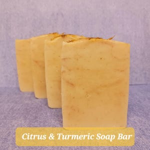 Peut inclure: Quatre savons rectangulaires au citron et au curcuma sont disposés en diagonale. Les savons sont jaune clair avec des marbrures brunes et quelques petits morceaux d'herbes séchées sur le dessus. Le texte "Citrus & Turmeric Soap Bar" est sur une étiquette jaune.