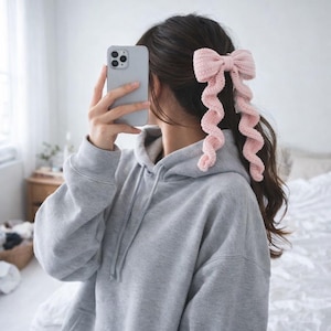 May include: A light pink, crocheted hair bow with two long, curly streamers. The bow is attached to the hair, and the streamers hang down. The person in the image is wearing a gray hoodie and holding a phone.