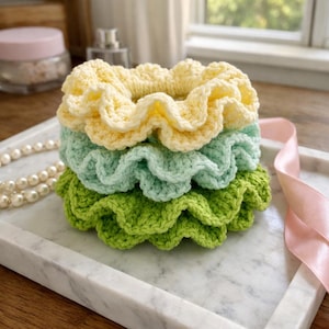 Könnte beinhalten: Ein Stapel von drei handgefertigten Häkel-Haargummis in Pastellfarben: Gelb, Mintgrün und Limettengrün. Die Haargummis haben einen gerüschten Rand und sind auf einem Marmortablett gestapelt. Eine Perlenkette und ein rosa Band sind ebenfalls zu sehen.