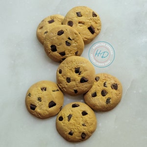 May include: Seven chocolate chip cookies arranged on a white marble surface. The cookies are round, with a golden-brown color and dark chocolate chips. The image includes a logo that says "HARPLINE DESIGNS ESTABLISHED 2010".