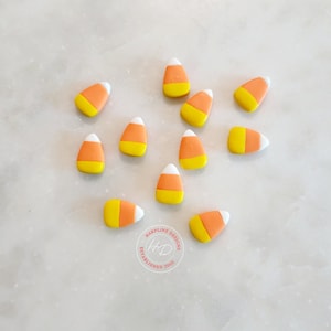 May include: A scattering of candy corn sweets, each with a white tip, orange centre, and yellow base. The sweets are arranged on a light-coloured surface. The image includes a logo for Harpline Designs, established in 2010.