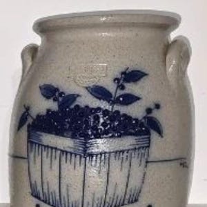 May include: A stoneware crock with a light beige glaze. The crock features a blue painted design of a basket filled with berries and leaves. It has two small handles on the sides.