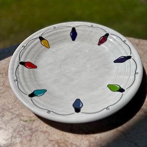 May include: White ceramic plate with a festive design of colorful Christmas lights. The plate features a circular arrangement of lights in various colors, connected by a gray line, creating a cheerful and decorative look. Perfect for holiday gatherings.