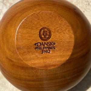 May include: A wooden bowl with a circular base. The bowl is a warm brown color and has a smooth, polished finish. The bottom of the bowl is stamped with the text "DANSK PHILIPPINES JHQ".