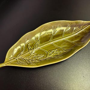 May include: A ceramic leaf-shaped dish in shades of green. The dish features detailed veining and leaf patterns, creating a naturalistic design. The elongated shape and glossy finish make it suitable for serving or display.