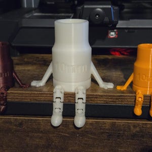 May include: Three 3D-printed figures resembling socket wrenches with arms and legs. The figures are in brown, white, and orange. Each figure has the text "10mm" printed on it. They are sitting on a wooden surface.
