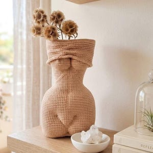 Puede incluir: Un jarrón de ganchillo beige con forma de torso, que contiene flores secas. El jarrón está sobre una estantería de madera con un cuenco blanco pequeño con cristales y una campana de cristal con una planta. El fondo incluye una ventana y cortinas transparentes.